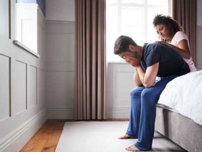 Woman Comforting Man Wearing Pajamas Suffering With Depression Sitting On Bed At Home