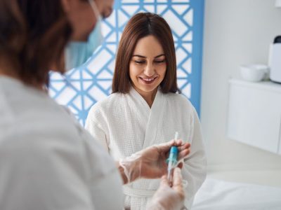 Beautiful woman in white bathrobe sitting on daybed and smiling while doctor holding syringe