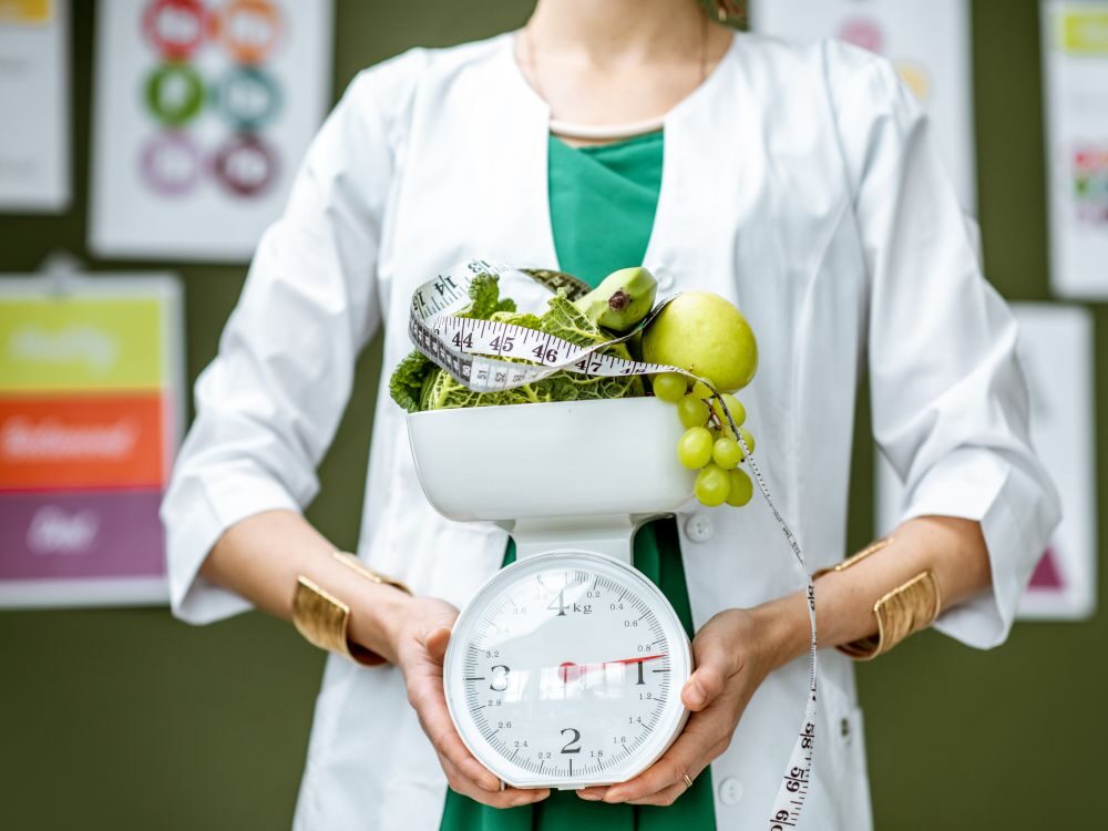 Young woman nutritionist in medical gown holding weights full of healthy products standing on the wall background with diet schemes